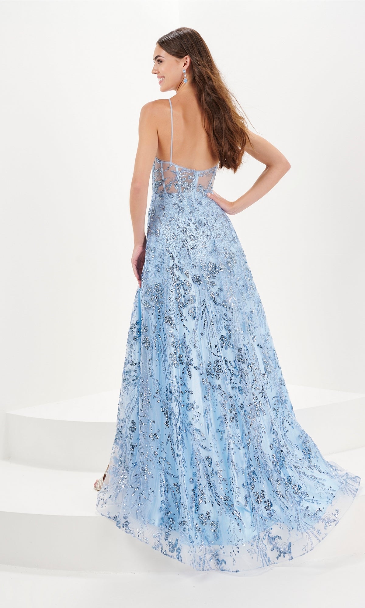 A woman stands on white steps, facing away, wearing the Tiffany A-Line Glitter Tulle Prom Dress 16097 in long light blue with silver floral patterns and a soft a-line silhouette.
